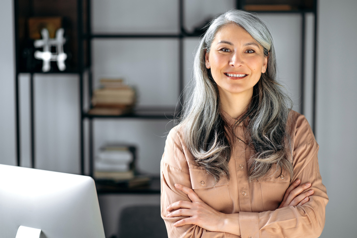 Portrait of a successful confident mature gray-haired lady, business woman, ceo or business tutor, standing in the office with arms crossed, looking and friendly smiling into the camera